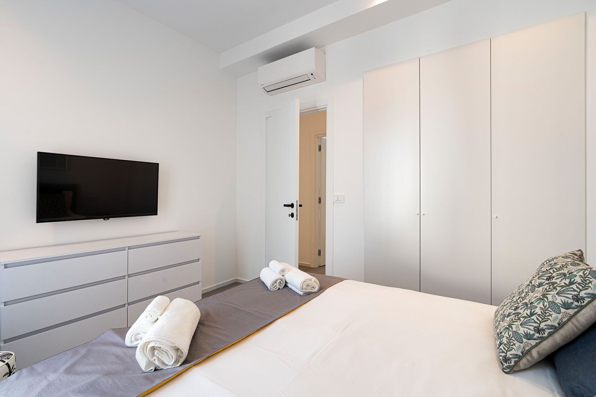 A modern hotel room with a flat-screen TV, a bed with a grey mattress, a white duvet, and a floral pillow. There are two rolled towels on the bed and a grey bedside table.