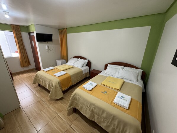 Habitación de hotel con dos camas, cada una con cubrecama color beige y almohadas blancas, y una mesita de noche pequeña con una lámpara.