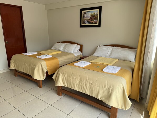 Une chambre d’hôtel avec deux lits, chacun recouvert d’une couvre-lit beige et agrémenté d’oreillers blancs. Un tableau encadré est accroché au mur au-dessus des lits, et une porte en bois se trouve sur le côté gauche.