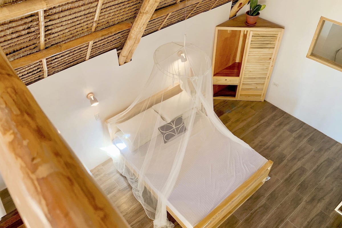 A room with a bed under a mosquito net, wooden flooring, a small wooden cabinet, and a potted plant on a table.