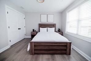 A bedroom with a wooden bed, white walls, a window with blinds, and a door. - Rest in Westerly (Westerly)