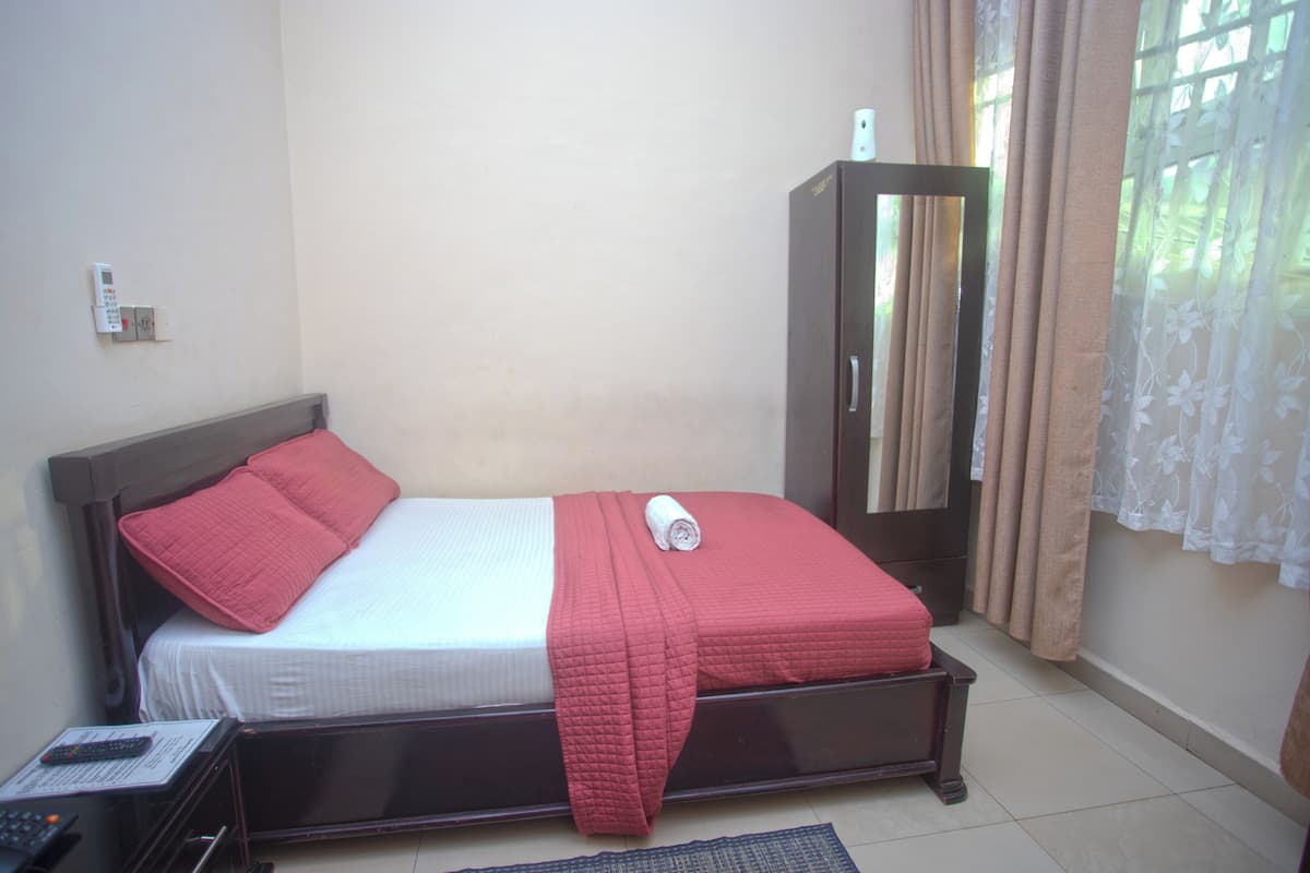 A neatly made bed with a red blanket and pillow, a wooden wardrobe, and a remote control on a nightstand.