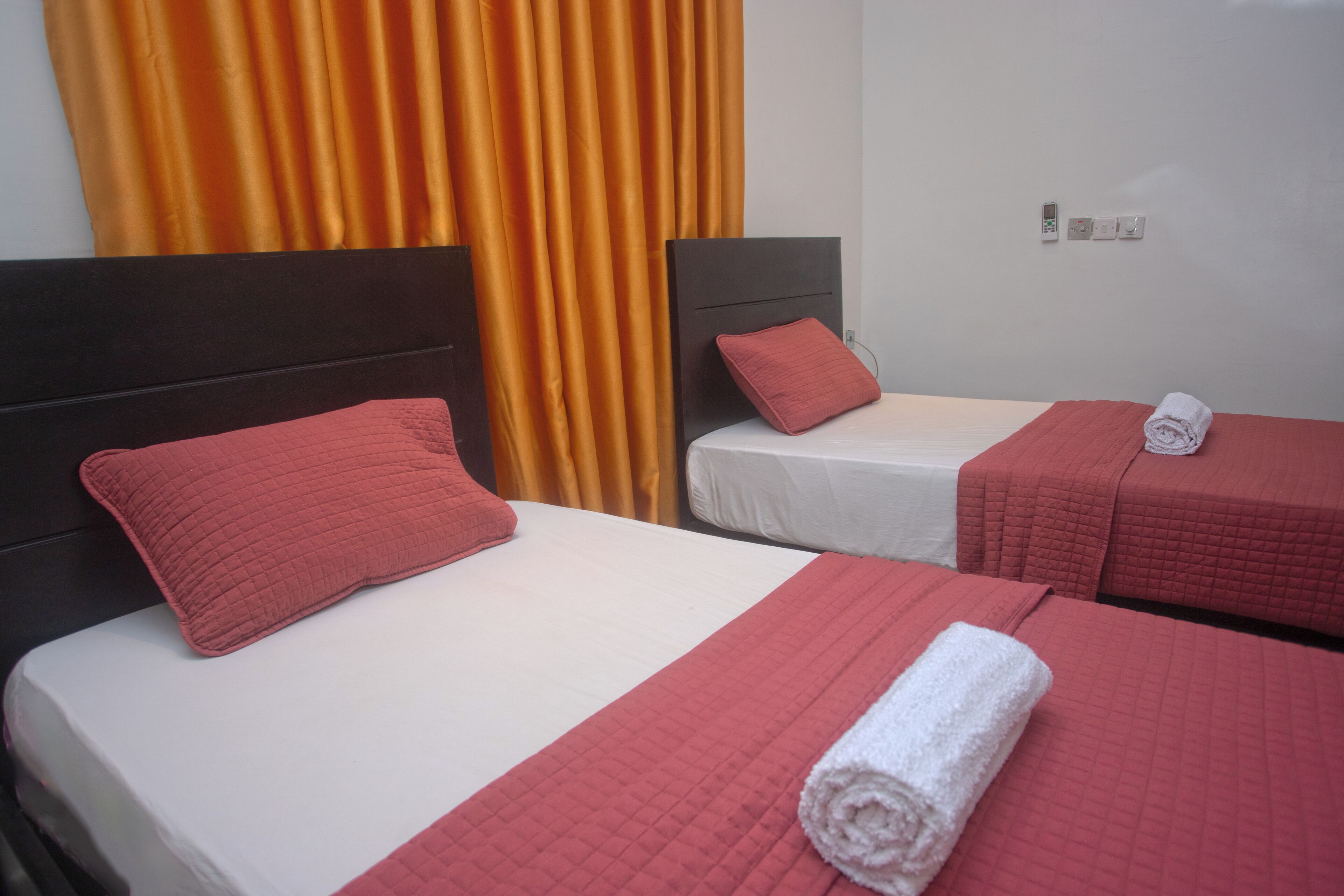 Two single beds with red bedding and pillows, a white wall, and an orange curtain.