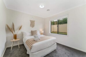 Un dormitorio con un ventanal grande, una cama con ropa de cama blanca, una mesita de noche con una lámpara y un tapiz de macramé en la pared.