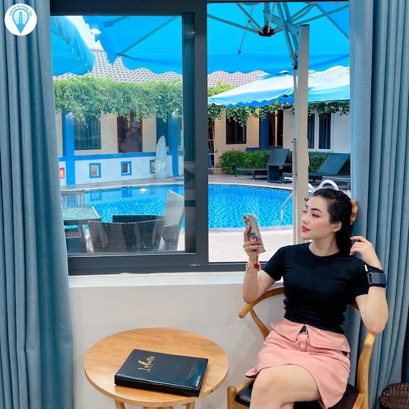 A person seated by a window overlooking a pool area with lounge chairs and a fountain.