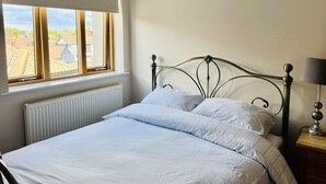 Een slaapkamer met een bed, een raam met uitzicht op gebouwen, een radiator en een nachtkastje met een lamp.