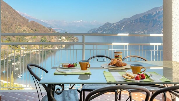 Mesa en un balcón con alimentos para el desayuno y una vista panorámica de un lago y montañas.