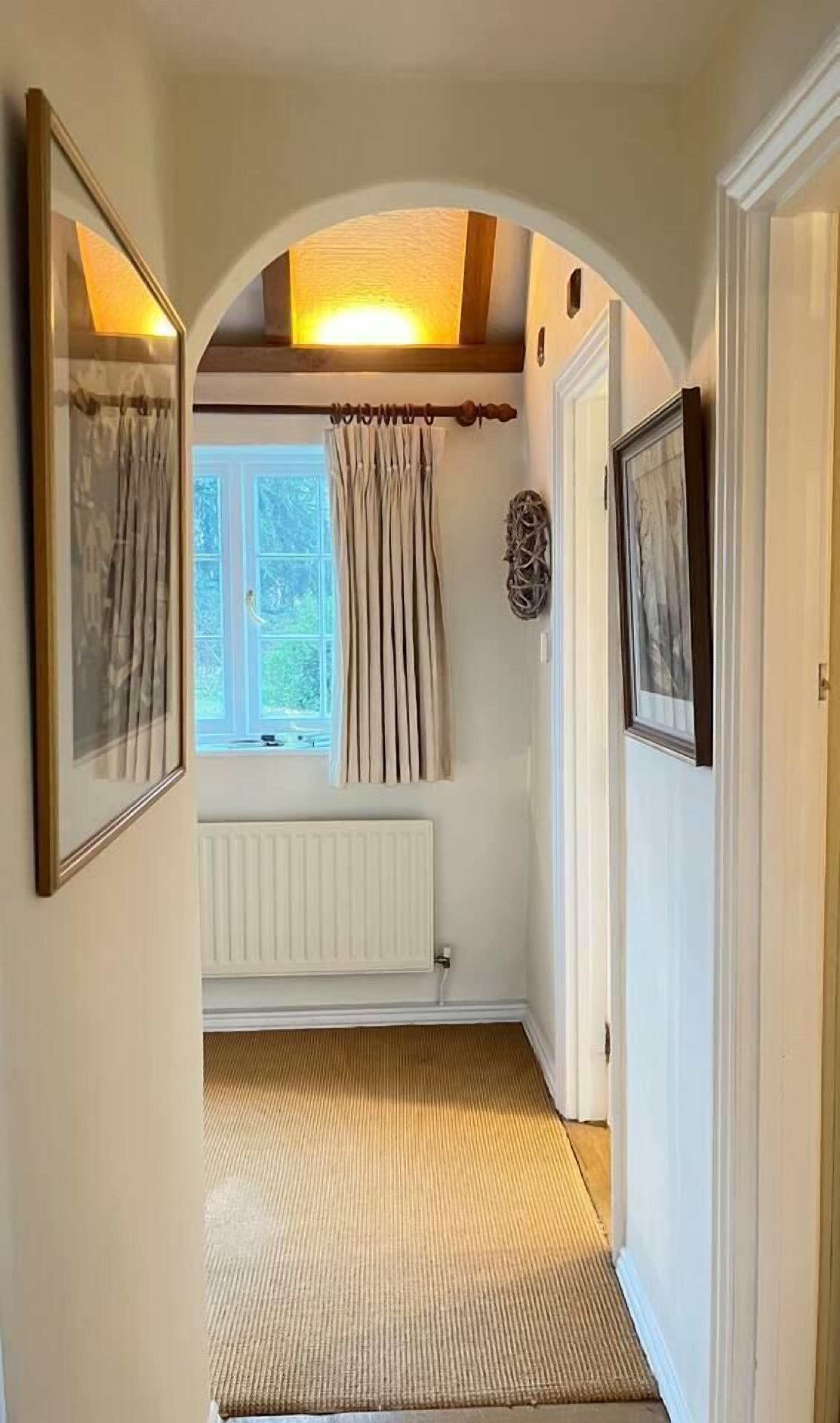 A narrow hallway with a radiator, a window with curtains, and framed pictures on the walls.