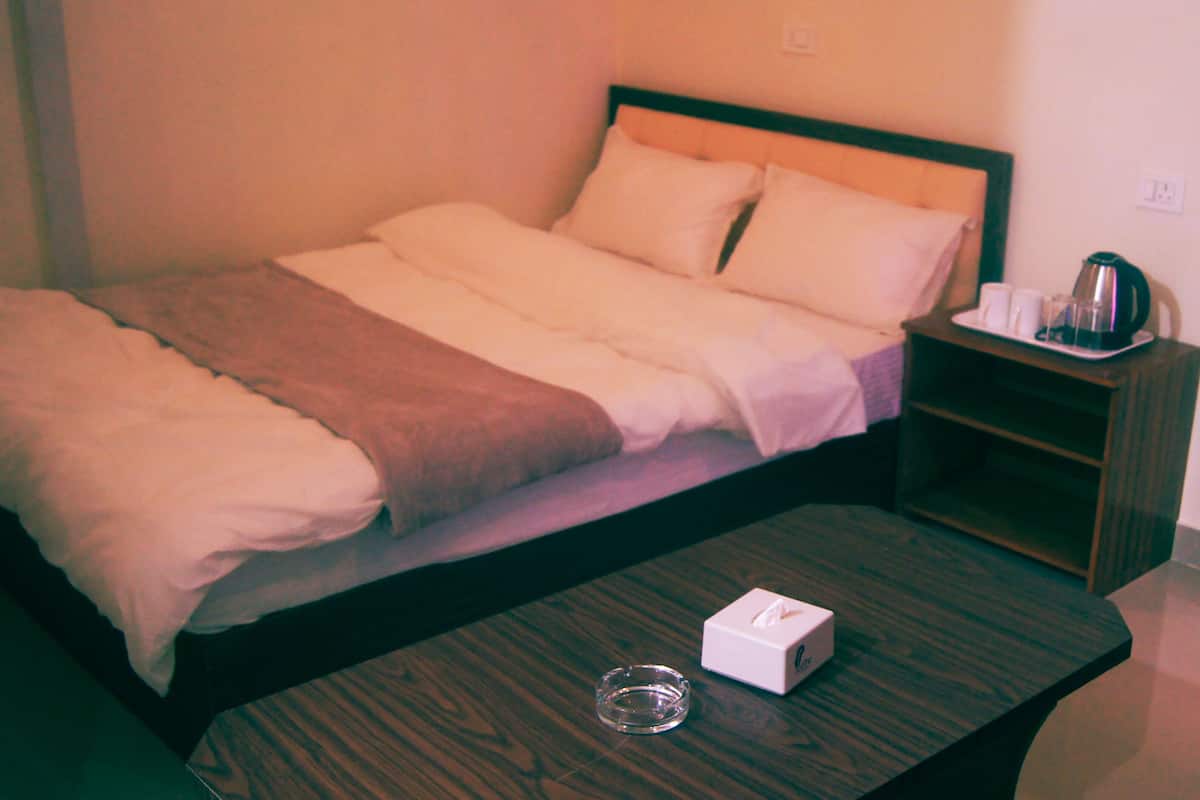 A neatly made bed with a brown blanket, a bedside table with a tissue box and a glass, and a wall-mounted light switch.