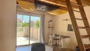 Une pièce dotée d’une porte-fenêtre coulissante donnant sur un balcon, d’une petite table avec une chaise, d’un fauteuil en forme de poire et d’une échelle.