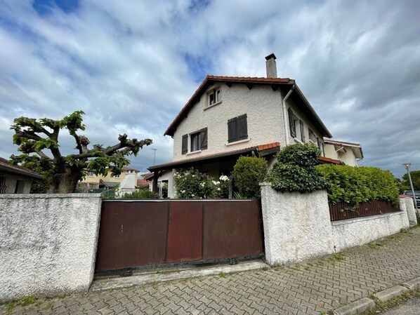 Una casa de dos pisos con un portón marrón, rodeada por un muro de piedra bajo y un seto.