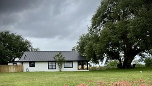 Una casa con techo inclinado, un pequeño jardín con flores rosadas y un árbol grande al fondo.