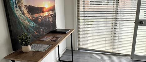 A room with a wooden desk, a large painting of a wave, a potted plant, and a sliding glass door with blinds.