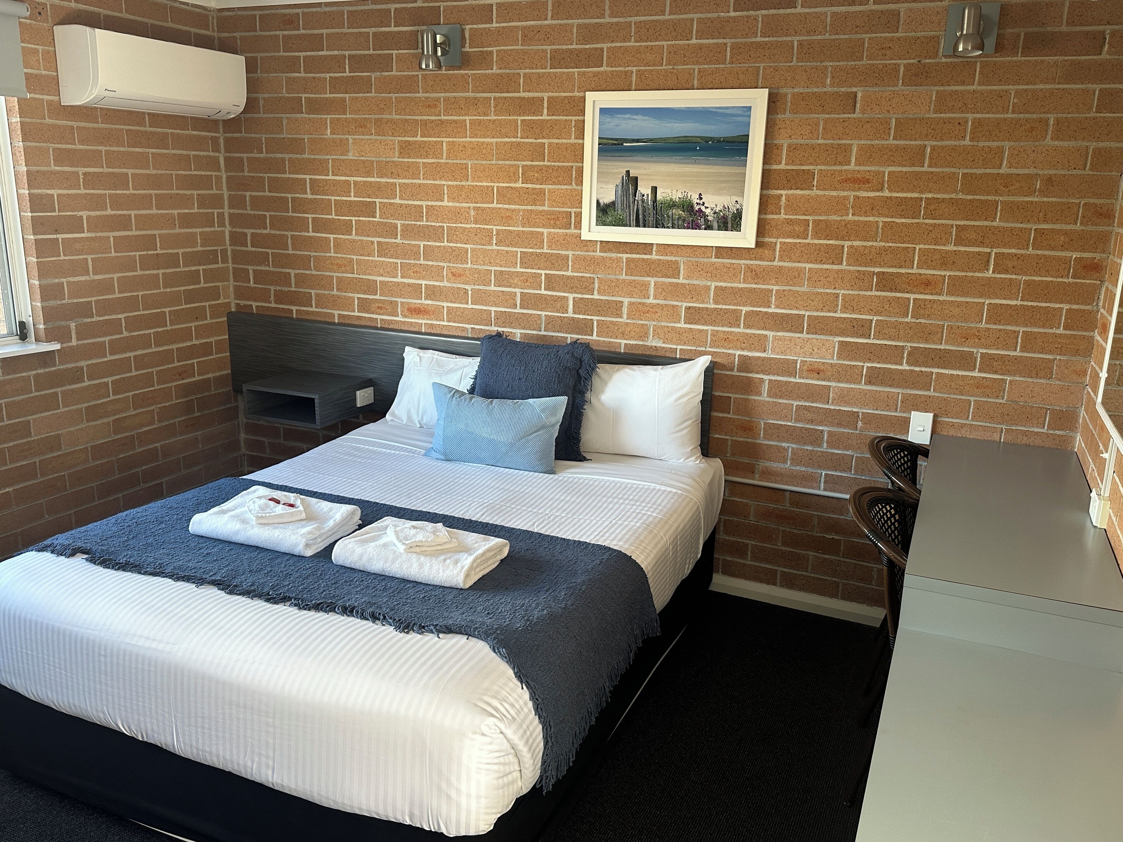 A hotel room with a brick wall, a bed with white and blue bedding, a framed picture on the wall, and a small desk with a chair.