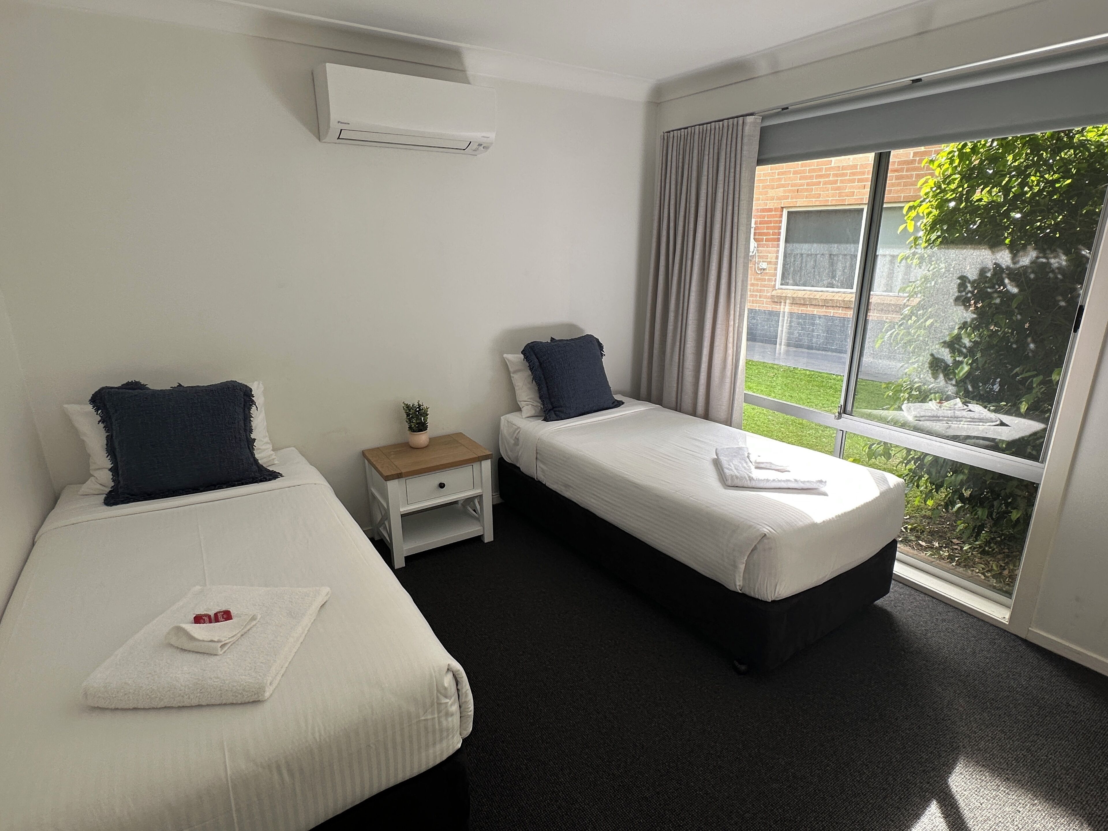 A hotel room with two beds, a small bedside table with a plant, a window with curtains, and an air conditioning unit on the wall.
