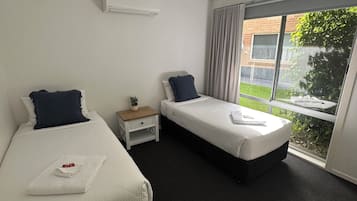 A hotel room with two beds, a small bedside table with a plant, a window with curtains, and an air conditioning unit on the wall.