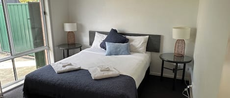 A hotel room with a large bed, two bedside tables with lamps, a window with a view of greenery, and an air conditioning unit on the wall.
