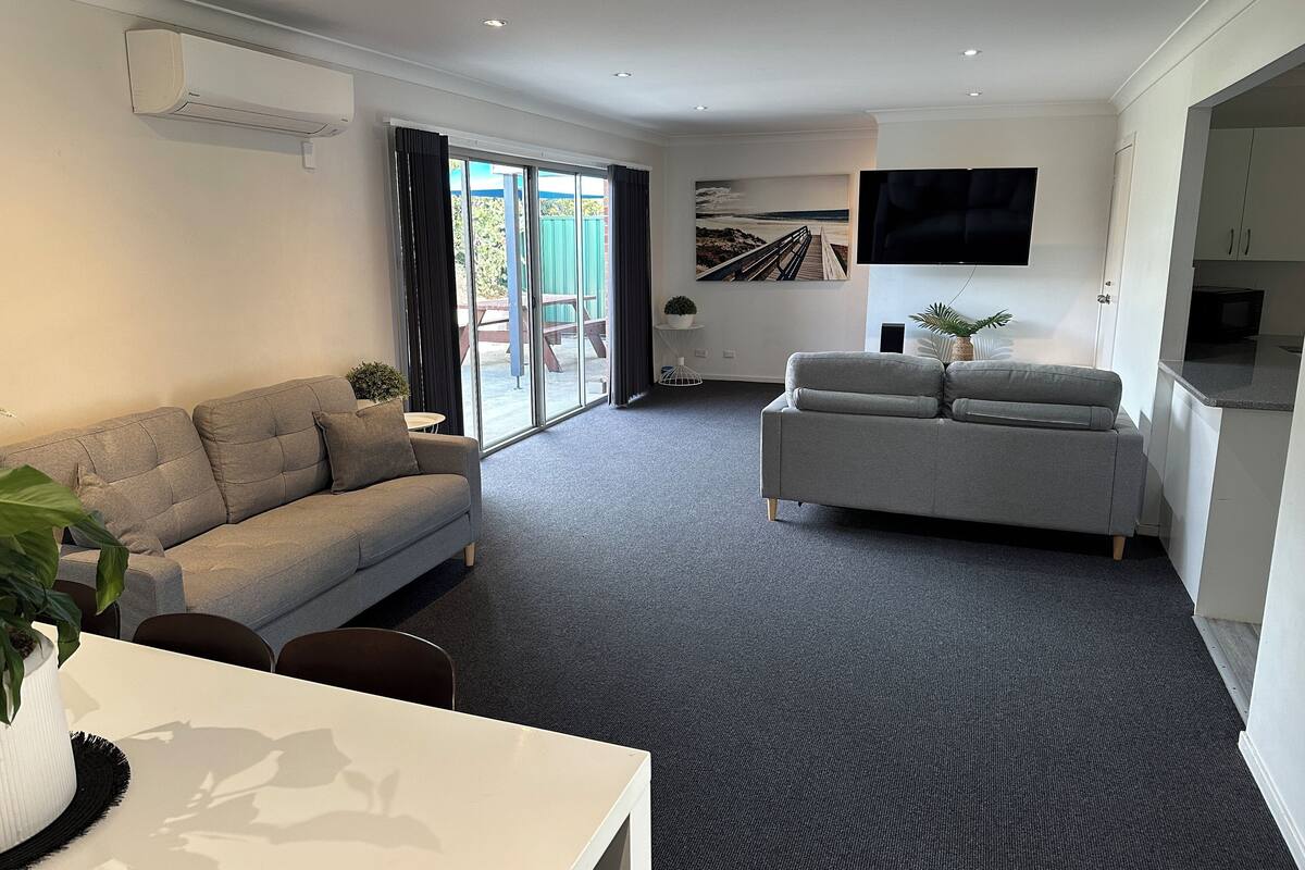 Ein modernes Wohnzimmer mit einem grauen Sofa, einem weißen Tisch mit Pflanze und einem grauen Teppich.