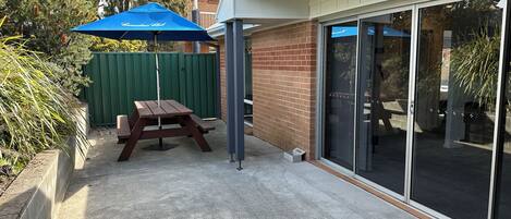 Un espace patio avec une table en bois et un parasol bleu, une clôture verte et un mur en briques.