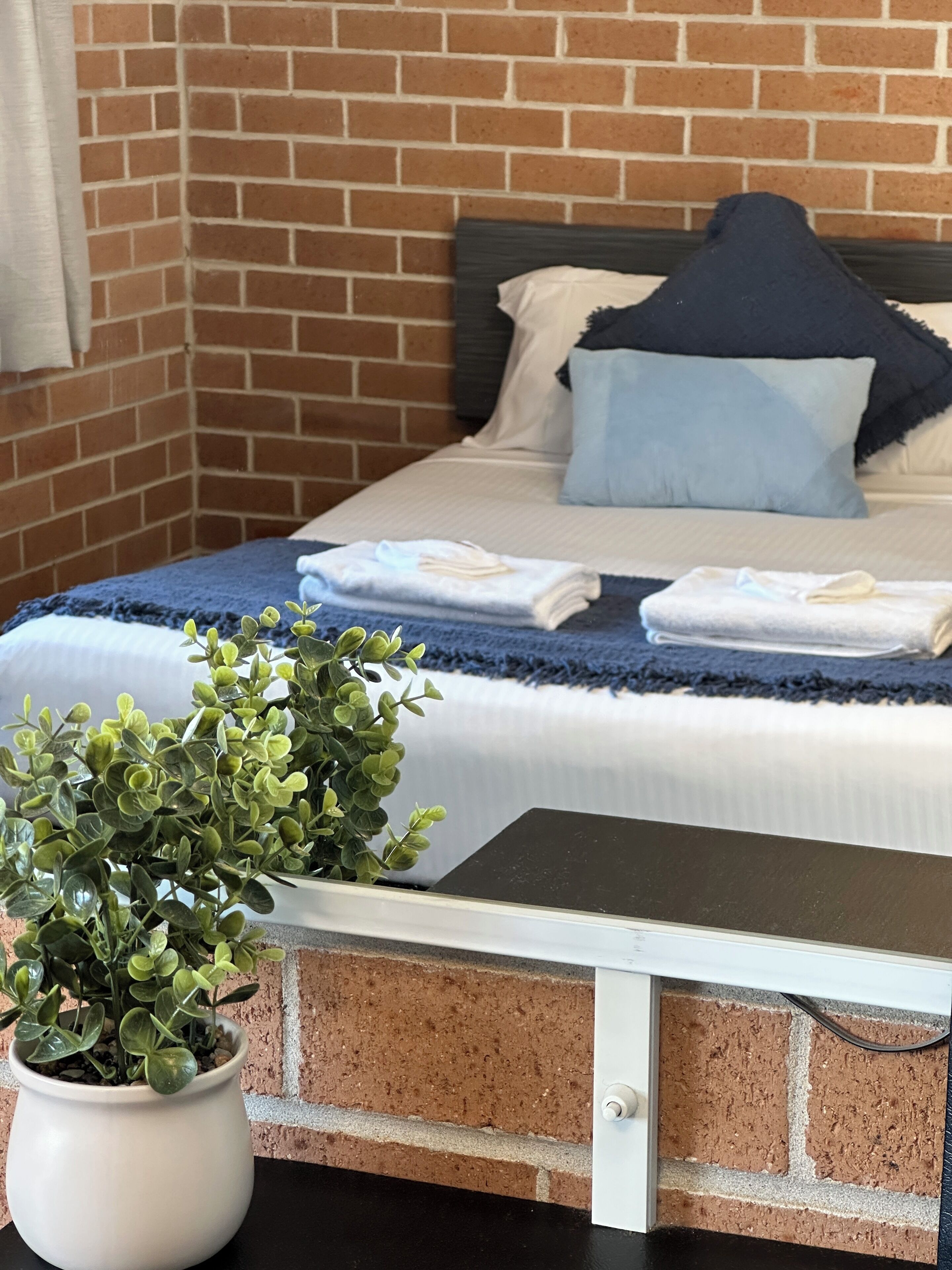 Habitación de hotel con pared de ladrillo, cama con ropa blanca y cabecero oscuro, manta azul y planta en maceta sobre la mesita de noche.