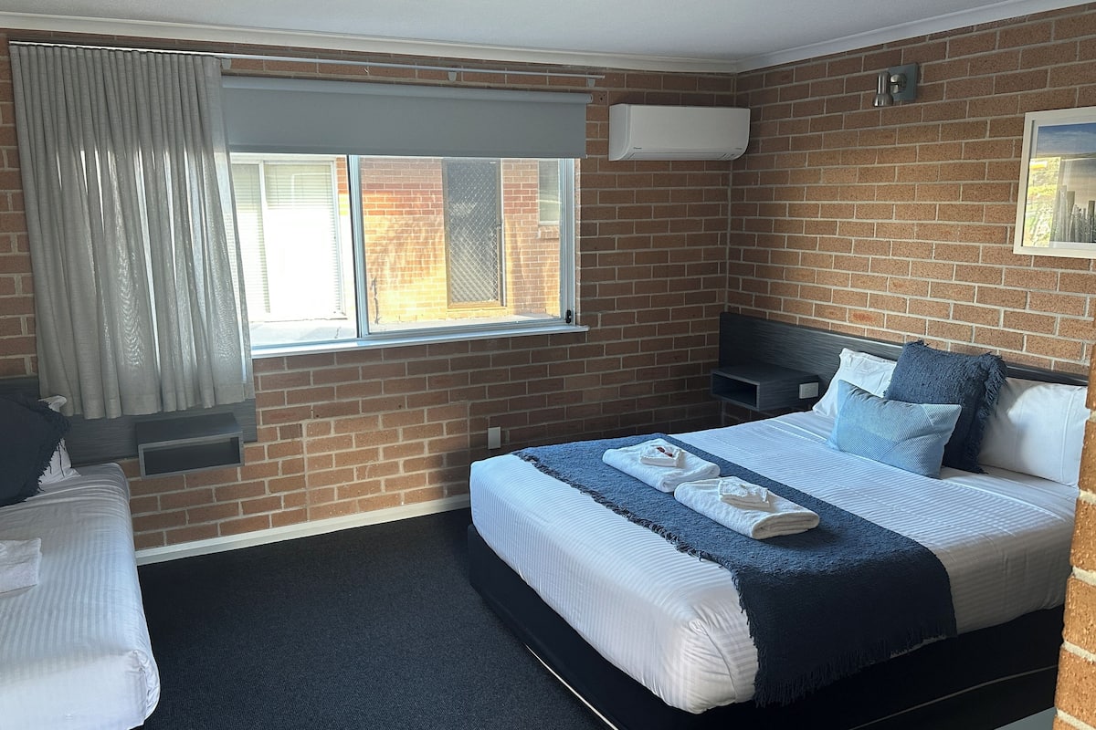 Ein Hotelzimmer mit Backsteinwand, einem Bett mit weißblauer Bettwäsche, einem Fenster mit Vorhängen und einem kleinen Nachttisch.