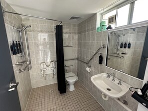 A modern bathroom with a shower, toilet, and sink. It includes grab bars, a hand dryer, and a mirror. - The Oaks Hotel Motel (Albion Park Rail)