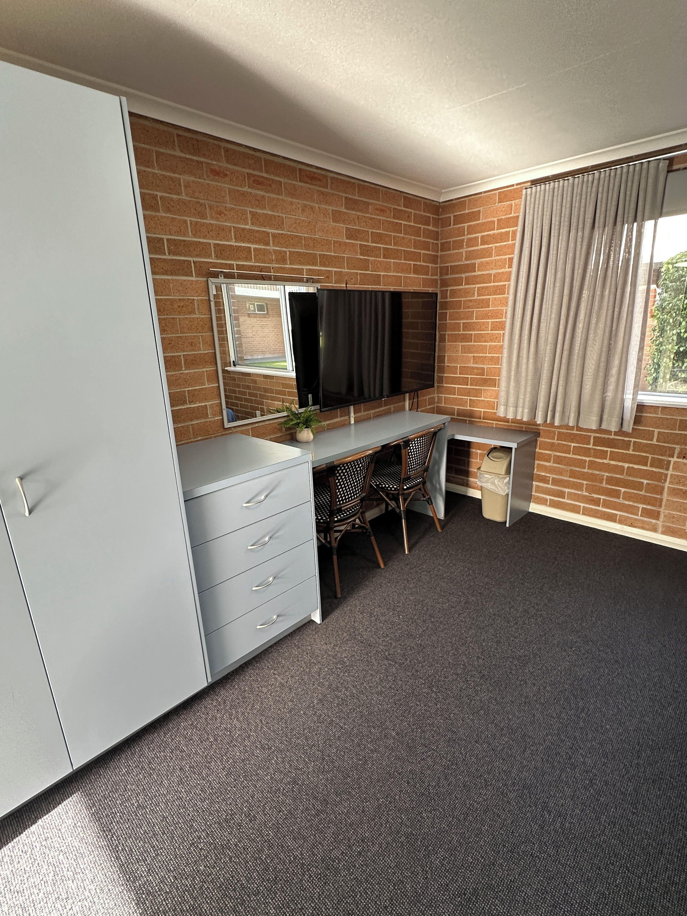 Ein Zimmer mit Backsteinwand, einem Wand-Fernseher, einem Schreibtisch mit zwei Stühlen, einem weißen Kleiderschrank und einem Fenster mit Vorhängen.