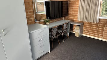 A room with a brick wall, a flat-screen TV mounted on the wall, a desk with two chairs, a white wardrobe, and a window with curtains.