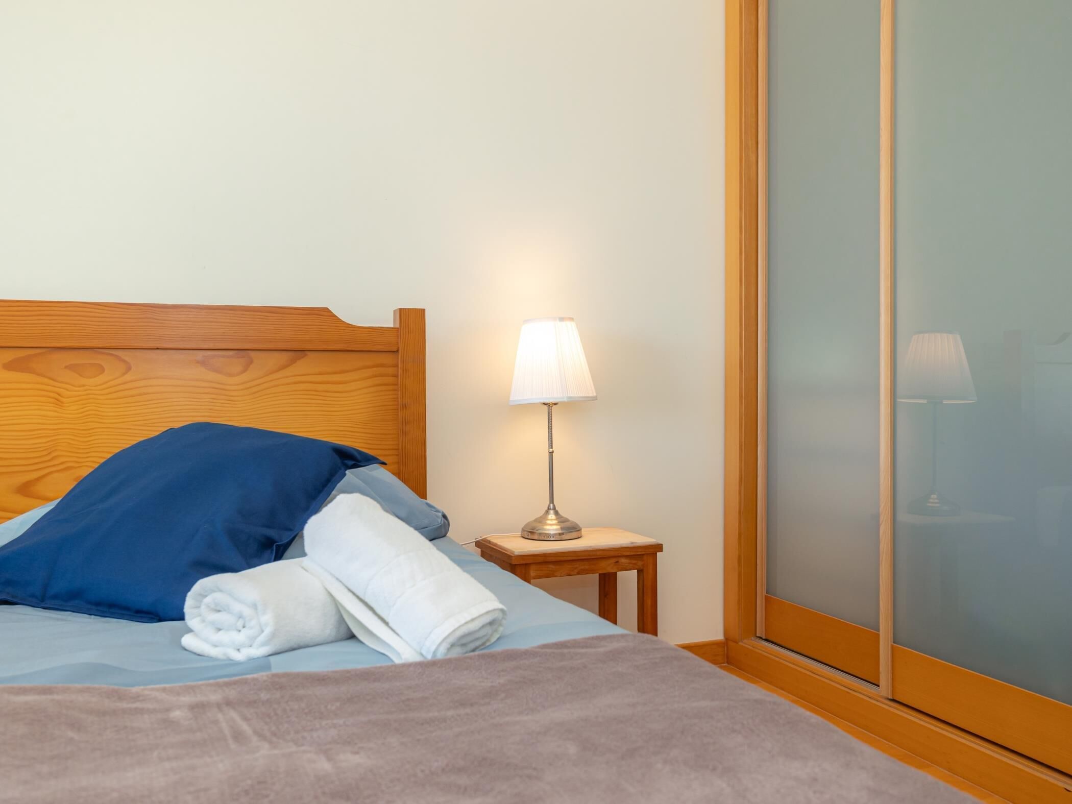 Ein ordentlich bezogenes Bett mit einem blauen Kissen und einem aufgerollten weißen Handtuch, ein kleiner Nachttisch mit einer Lampe sowie eine Schiebetür aus Glas.