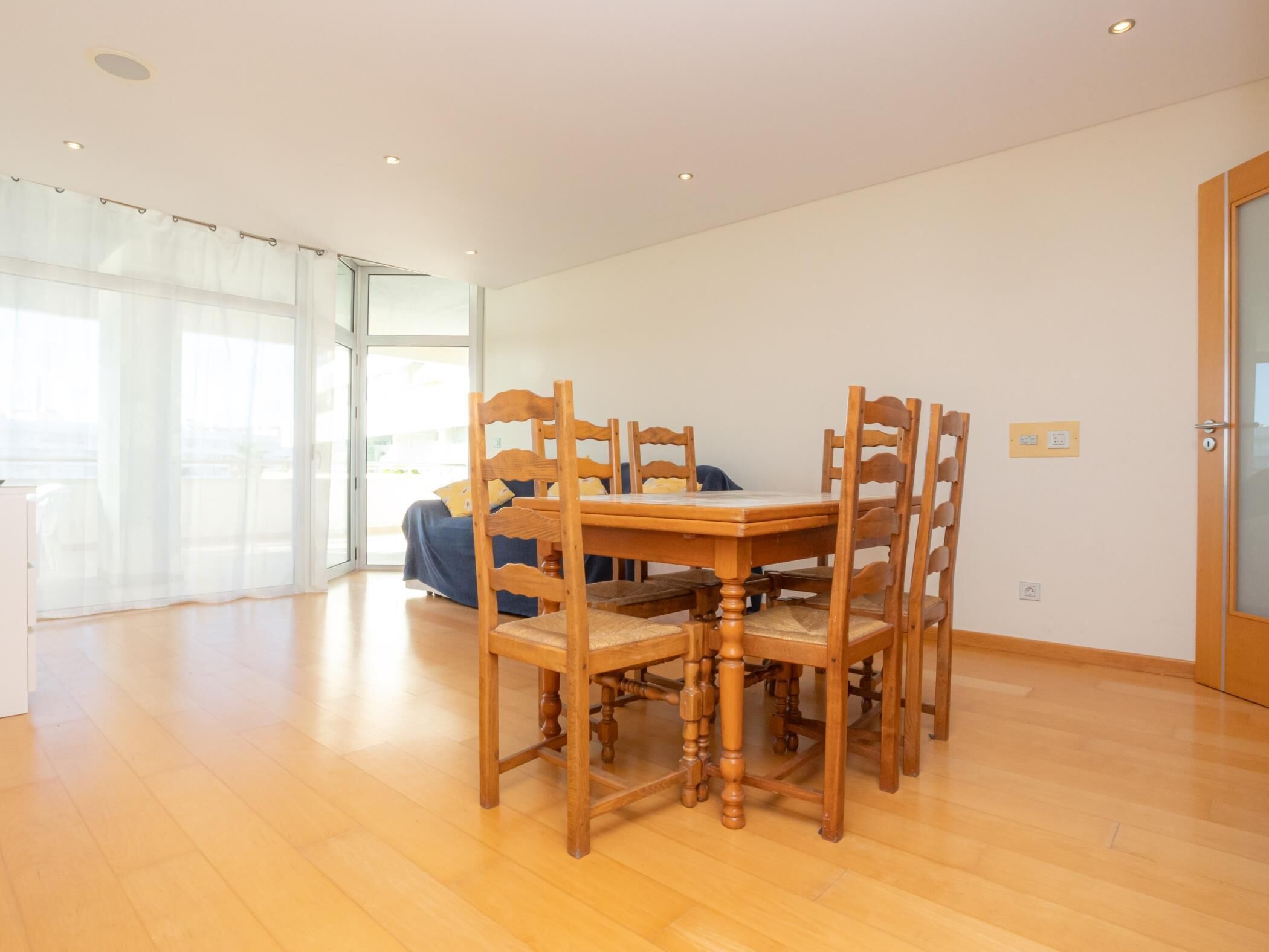 A dining area with a wooden table and chairs, a bed with a blue bedspread, and a large window with sheer curtains.
