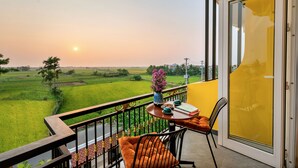 A balcony with a view of green fields and a sunset, a table with a vase of flowers, and a chair.