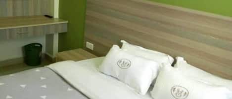 Una cama bien hecha con colcha gris y almohadas blancas, cabecera de madera, pared verde y televisor montado en la pared.