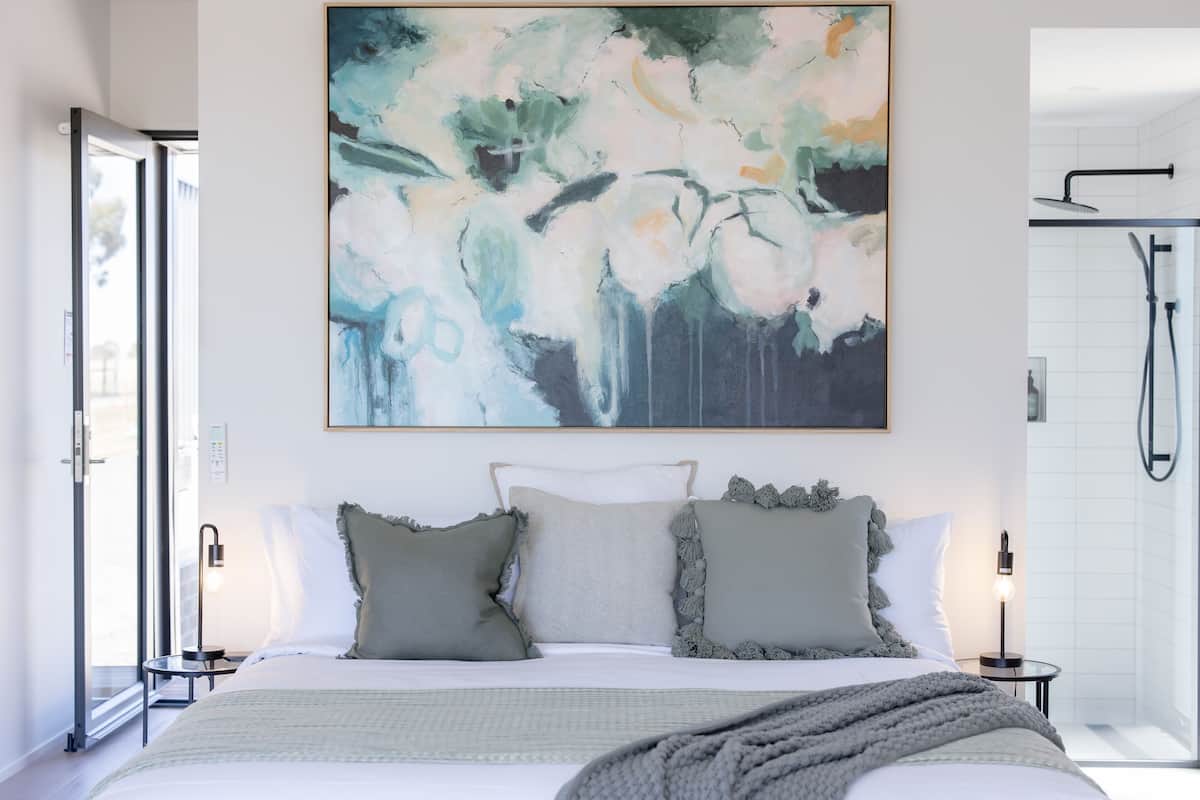 A modern bedroom with a large abstract painting, a bed with grey and white bedding, and a visible shower area.