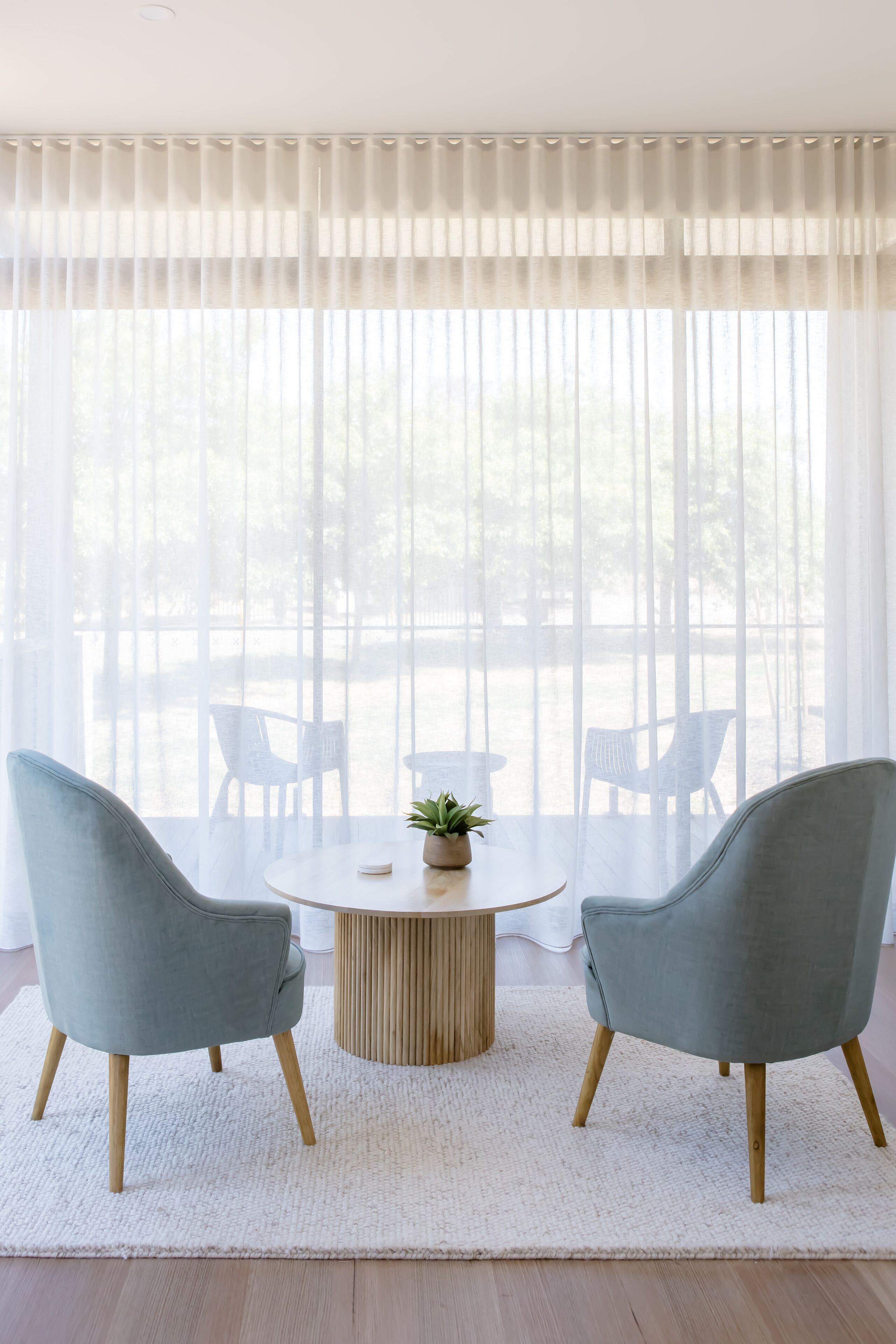 Una habitación con una mesa redonda, dos sillas, una planta y vistas a través de cortinas de gasa.