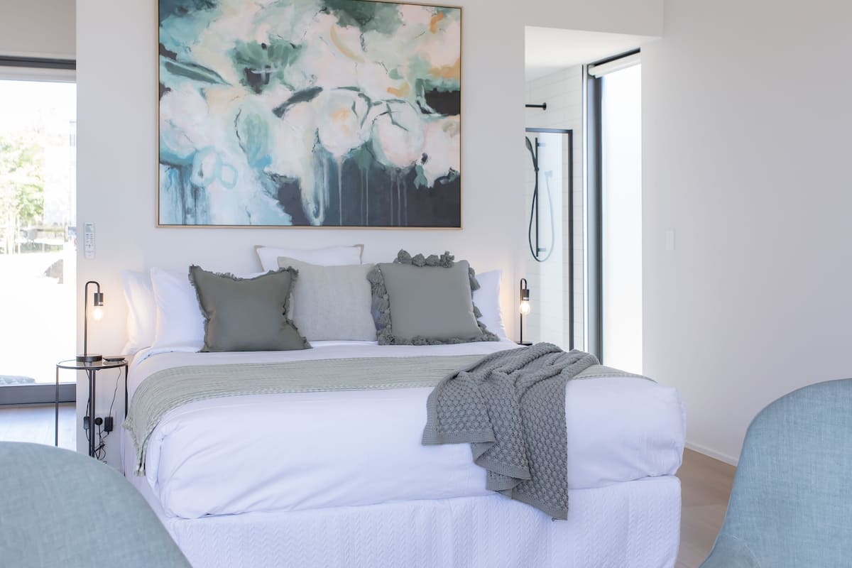 A modern bedroom with a large bed, two armchairs, a round coffee table, and a large abstract painting on the wall.