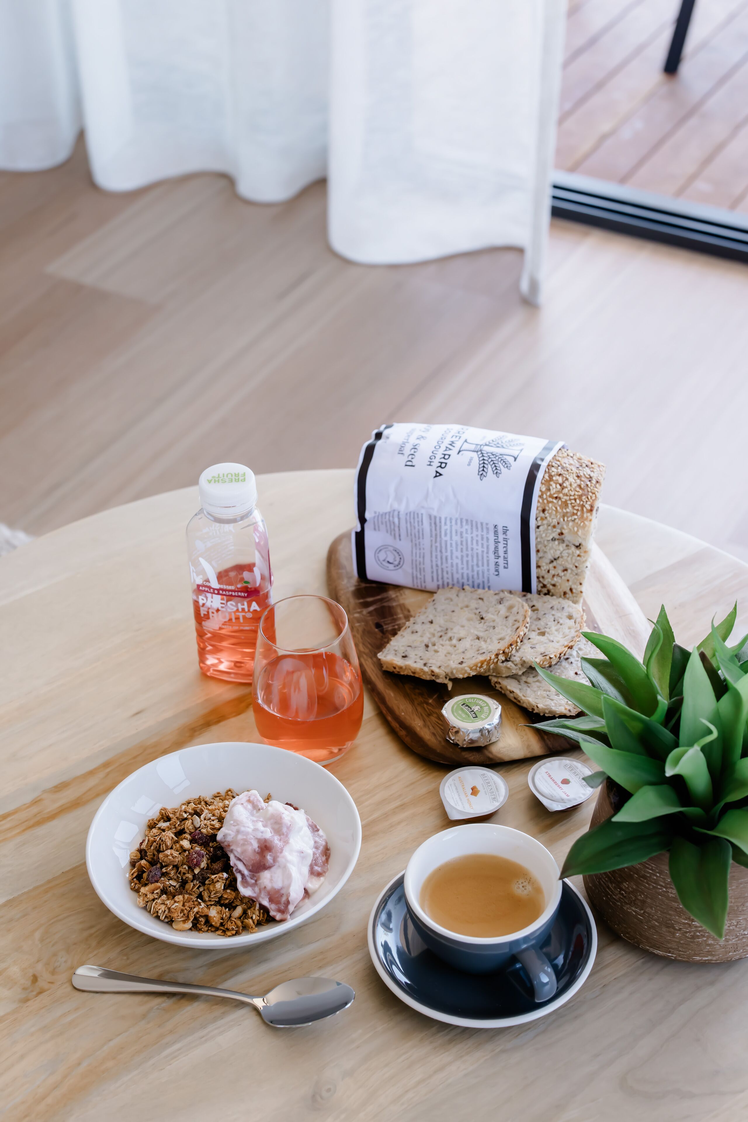 A breakfast setting with a bowl of granola and yogurt, a cup of coffee, a bottle of juice, and a loaf of bread on a wooden table.
