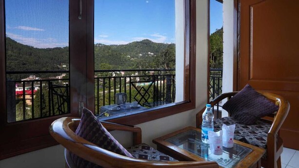 A balcony with a view of a green landscape, a table with a bottle, and a chair. - Hotel Apple Inn (Nainital)
