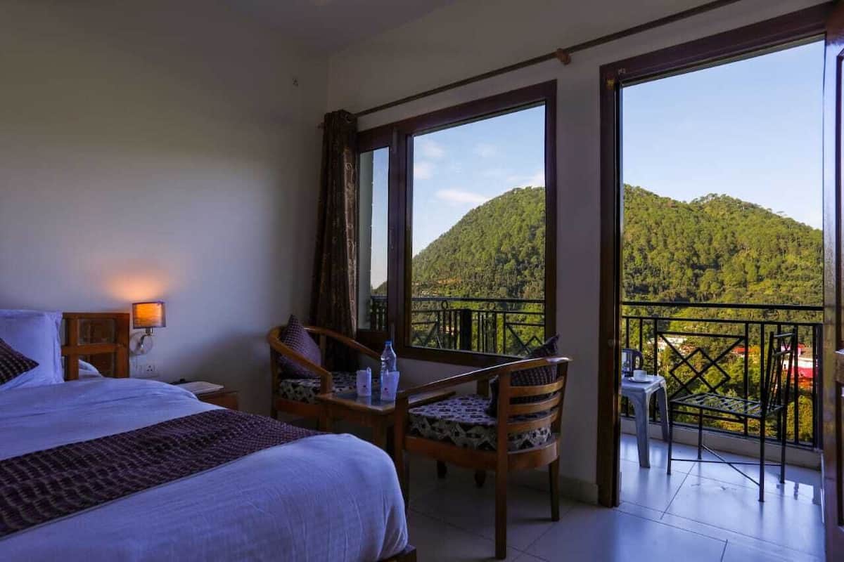 A hotel room with a bed, a table with chairs, and a balcony with a view of a forested hill.