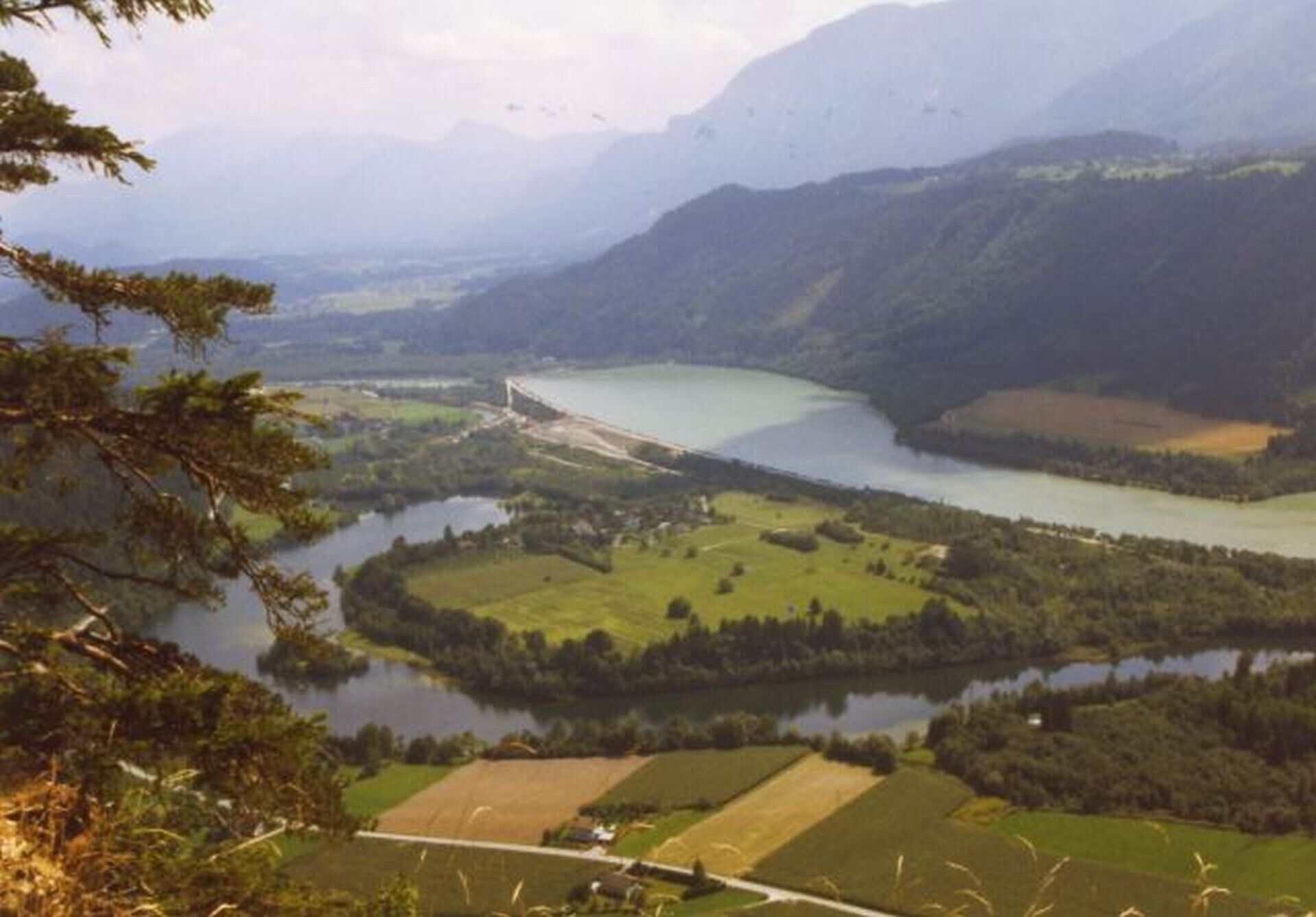 Een bergachtig landschap met een rivier, velden en een brug.
