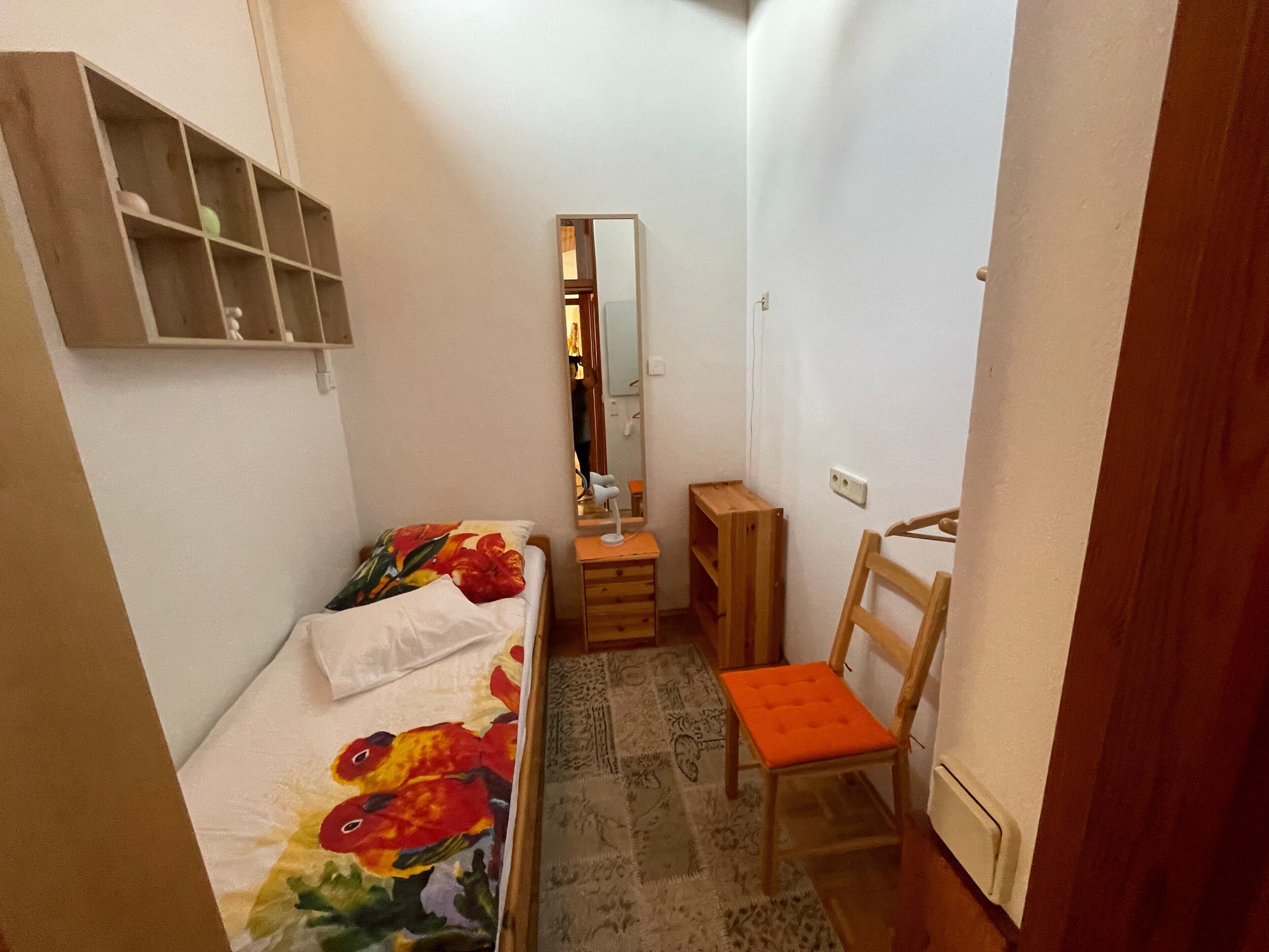 A small bedroom with a bed, a chair, a dresser, a mirror, and a shelf.