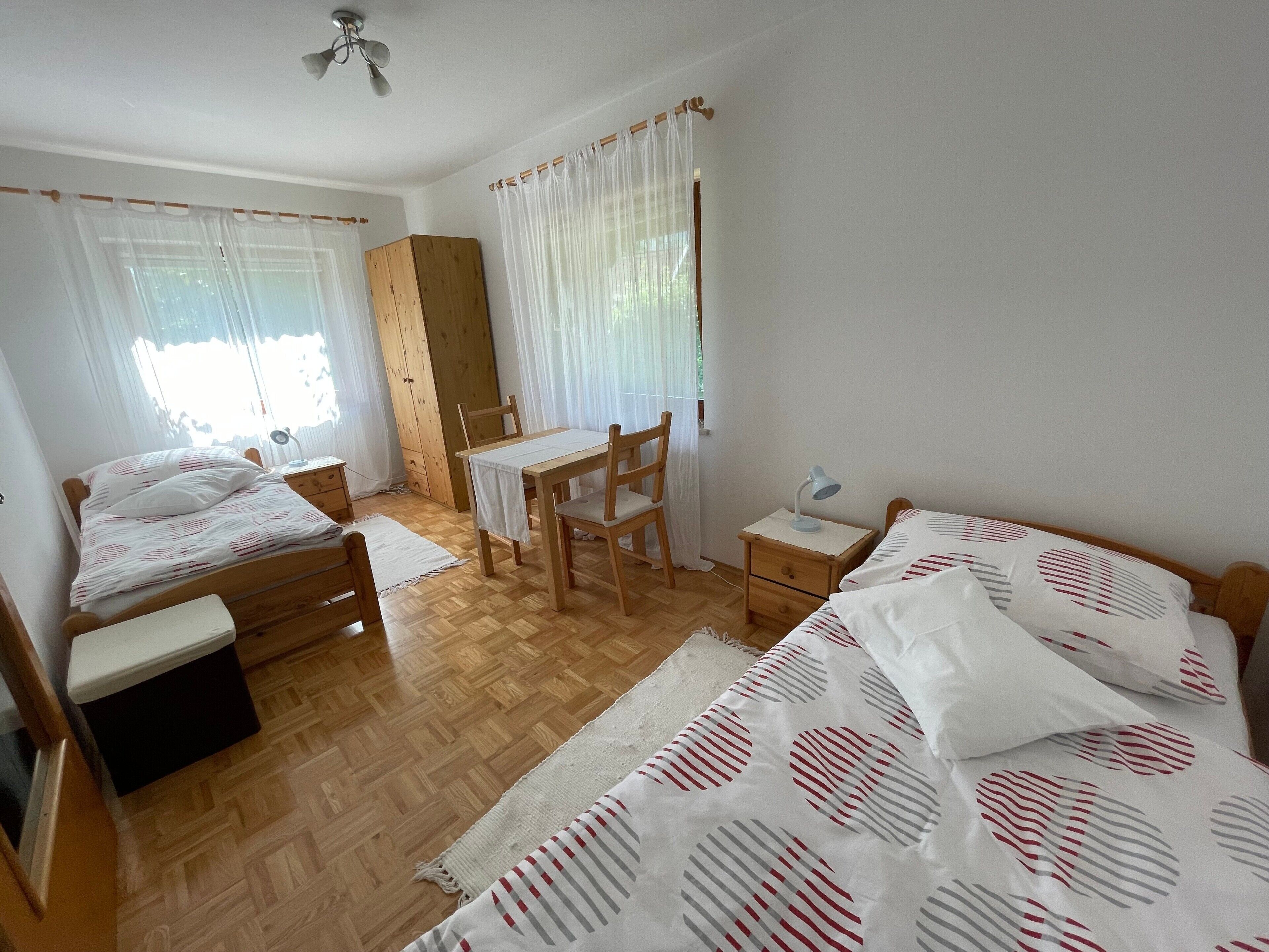 A bedroom with two beds, a wooden wardrobe, a desk with a chair, and a nightstand with a lamp.