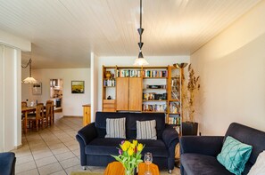 Een woonkamer met een bank, een bijzettafeltje met bloemen, een boekenkast en een eethoek met stoelen en een tafel.