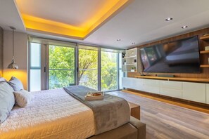 Quarto moderno com uma cama grande, uma TV fixada na parede e vista para árvores através de janelas amplas.