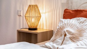 Een nachtkastje met een lamp, een bed met een wit kussen, en een lichtschakelaar aan de muur.
