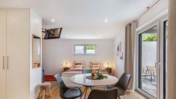 Een moderne kamer met een ronde eettafel, stoelen, een klein krukje, een bed met kussens, een raam en een schuifpui.