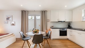 Een moderne keuken met witte kasten, een eethoek met een ronde tafel en stoelen, en een groot raam met gordijnen.