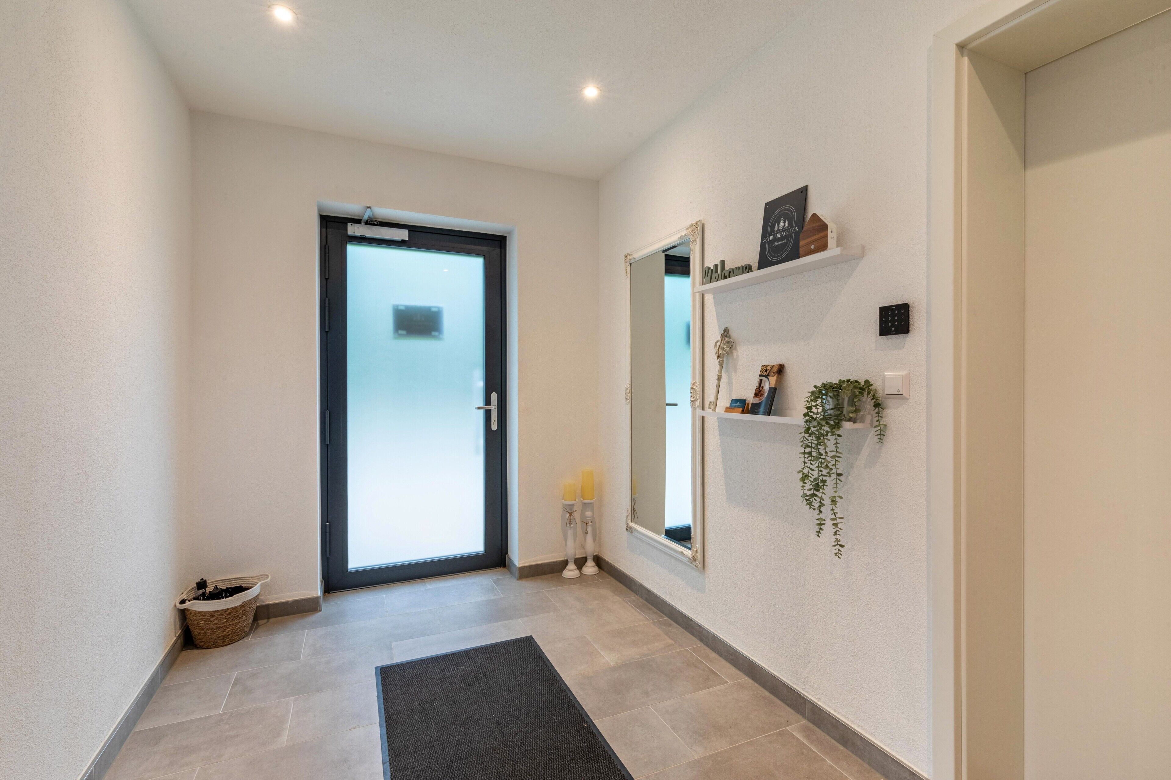 A hallway with a door, a mirror, a shelf with items, and a mat on the floor.