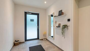 A hallway with a door, a mirror, a shelf with items, and a mat on the floor.