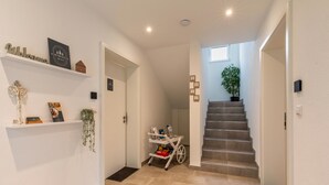 A hallway with a staircase, a shelf with decorative items, a plant, and a cart with books.