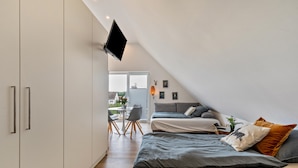 Una camera da letto con televisione a muro, un letto con biancheria grigia, un tavolino con sedie e un balcone con vista.